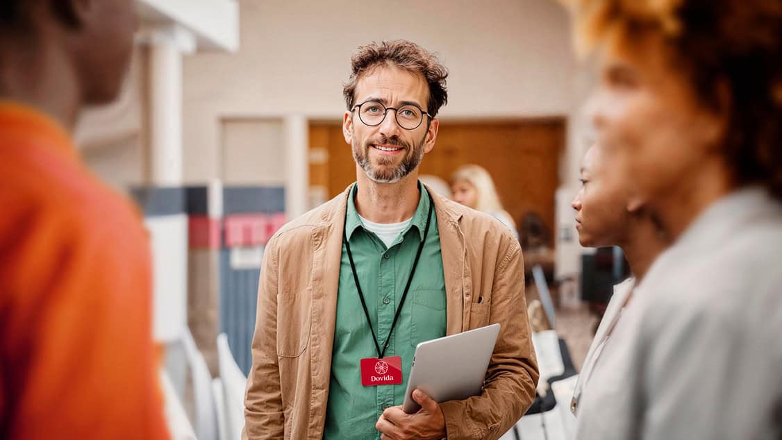 Dovida agent wearing glasses and carrying a laptop talks with colleagues at an event. He is wearing a name tag with the Dovida logo.
